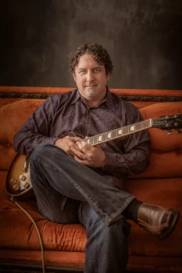 A man sitting on an orange couch with an electric guitar for his headshot.