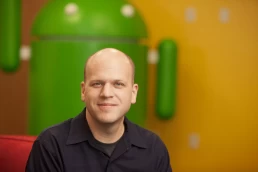 A bald man sitting in front of an android logo for a company headshot.