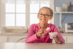 A girl with glasses is eating yogurt at a table in a food photography shoot.