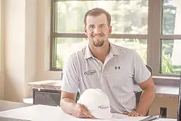 A man sitting at a desk with a hard hat, posing for a company headshot.
