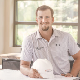 A man sitting at a desk with a hard hat, posing for a company headshot.