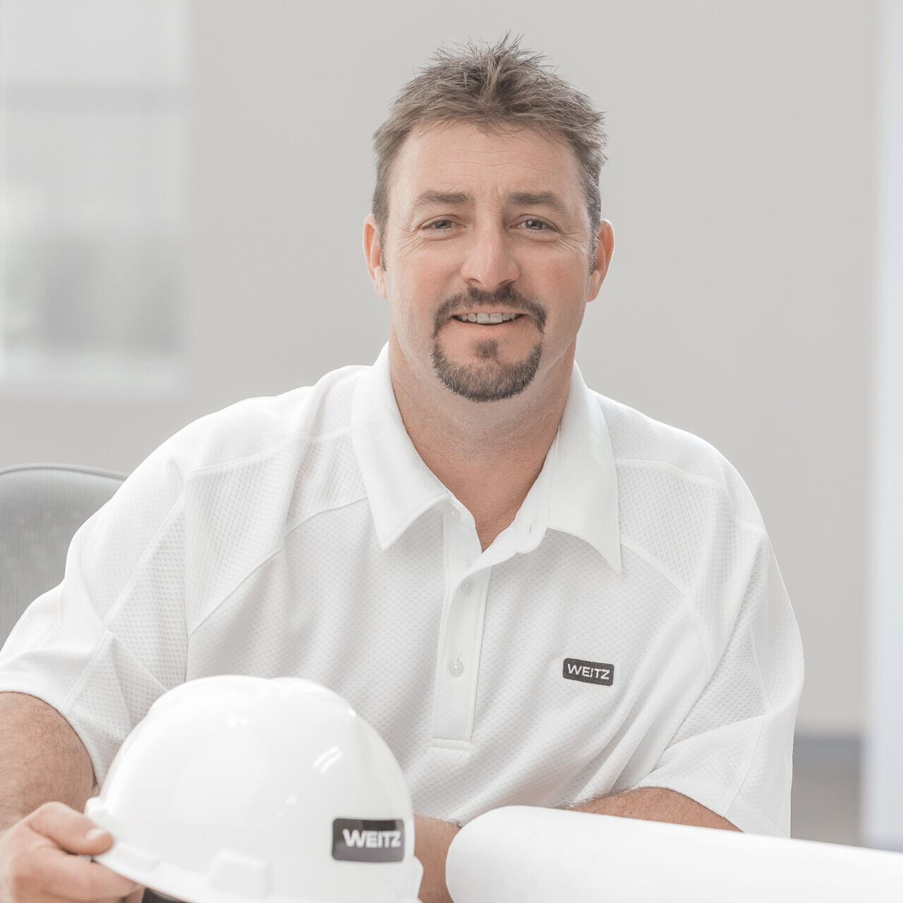 A man wearing a white hard hat and holding a company headshot.