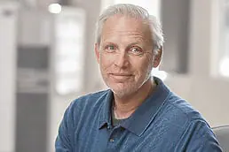 A man in a blue shirt sitting in a chair for a company headshot.