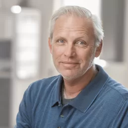 A man in a blue shirt sitting in a chair for a company headshot.