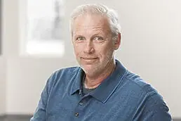 A man in a blue shirt is sitting in an office for his company headshot.