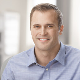 A smiling man in a blue shirt sitting at a desk for his company headshot.