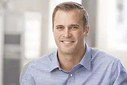 A smiling man in a blue shirt sitting at a desk for his company headshot.