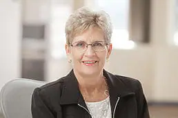 A woman wearing glasses is smiling in her headshot in an office.