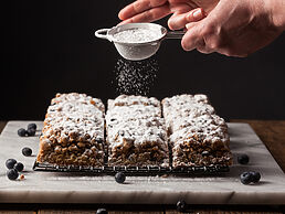 A person is sprinkling powdered sugar on a stack of blueberry bars.