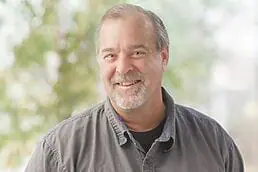 A man in a gray shirt smiling in front of trees for his company headshot.