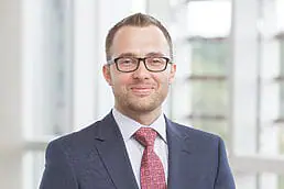 A man in a suit and tie standing in front of a window for a headshot.