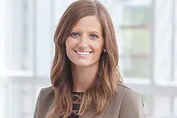 A woman in a blazer smiling in front of a window for her company headshot.