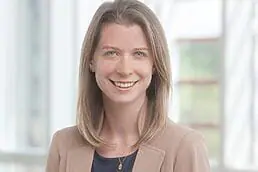 A woman in a tan blazer smiles in front of a window for her company headshot.