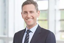 A man in a suit and tie smiling in front of a window for his headshot.