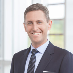 A man in a suit and tie smiling in front of a window for his headshot.