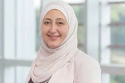 A woman in a hijab smiling in front of a window for her company headshot.