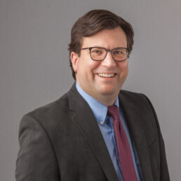 A man in a suit and tie smiling for the camera in his company headshot.