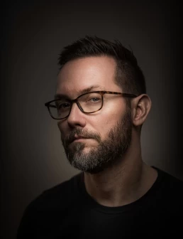 A company headshot of a man with glasses and a beard in front of a dark background.
