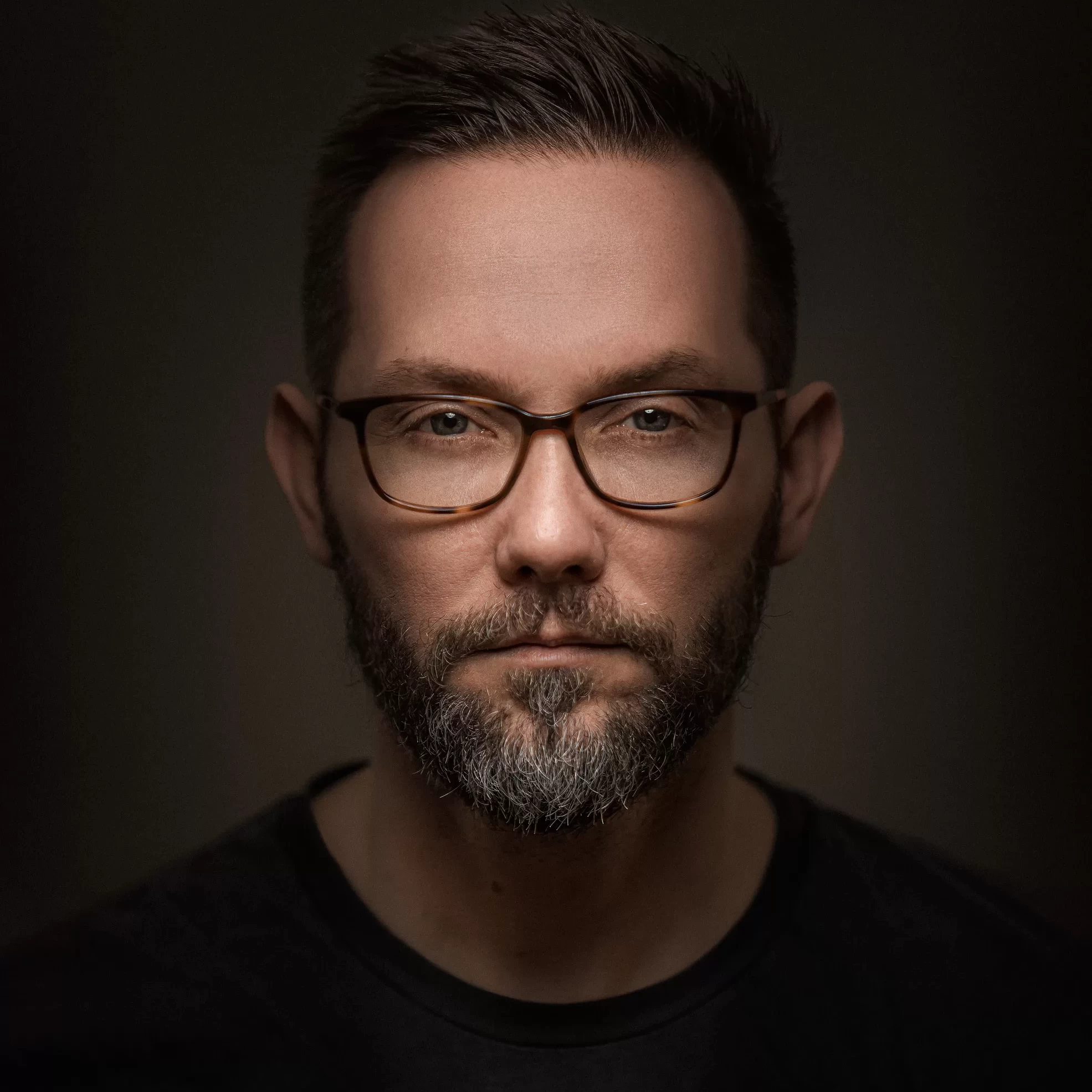 A man with glasses and a beard in front of a dark background for a company headshot.