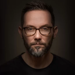 A man with glasses and a beard in front of a dark background for a company headshot.