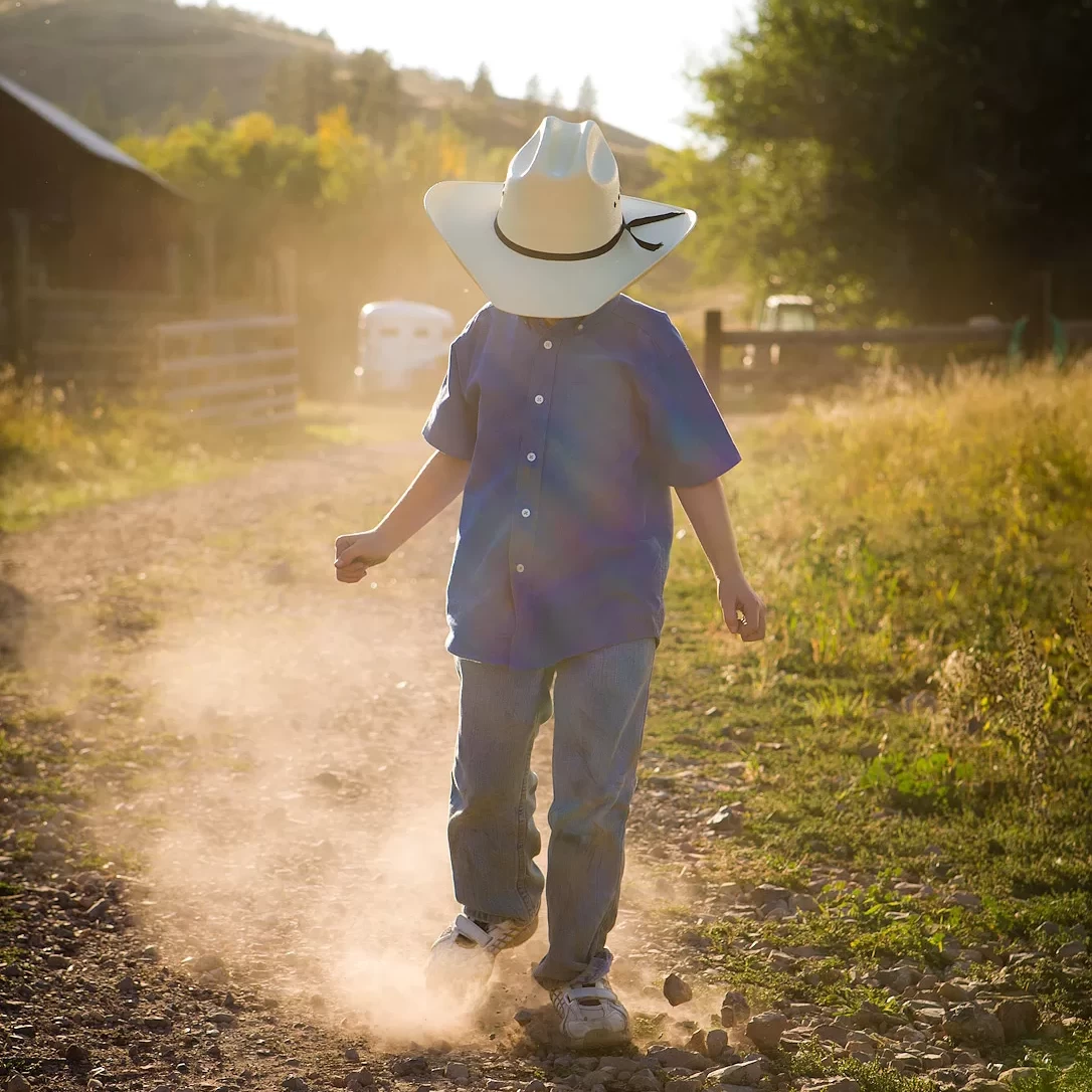 A boy wearing a cowboy hat walking down a dirt road photography shoot.