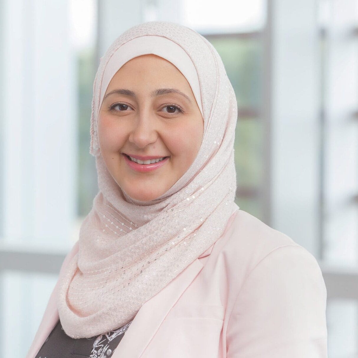 A woman in a hijab smiling in front of a window for her company headshot.