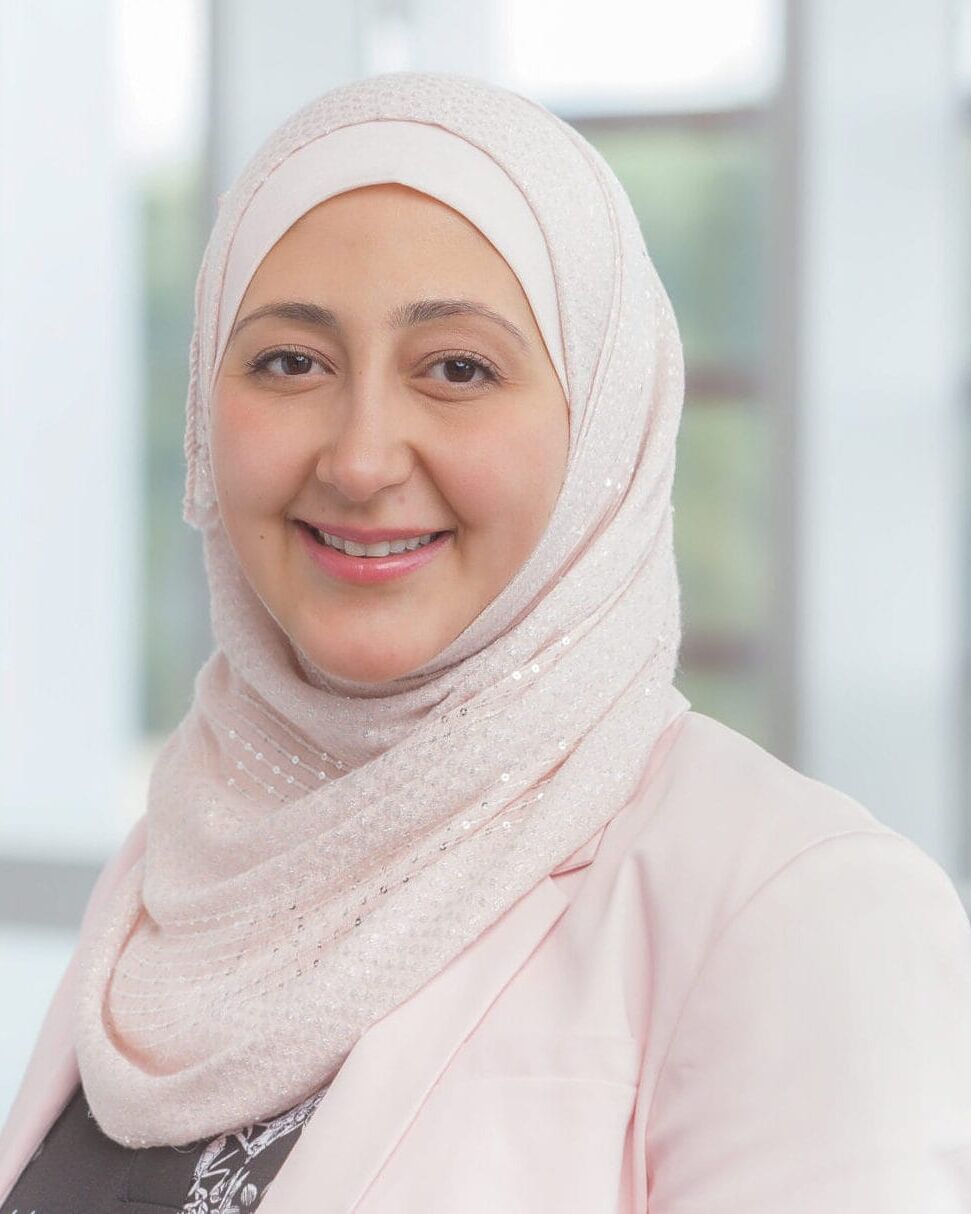 A woman in a hijab smiling in front of a window for her company headshot.