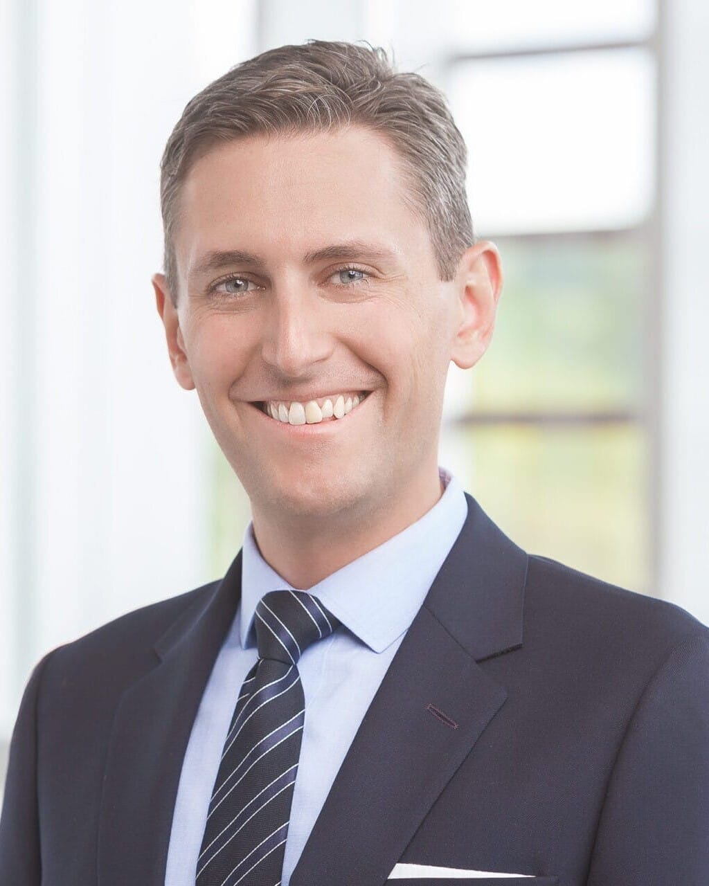 A man in a suit and tie smiling in front of a window for his headshot.