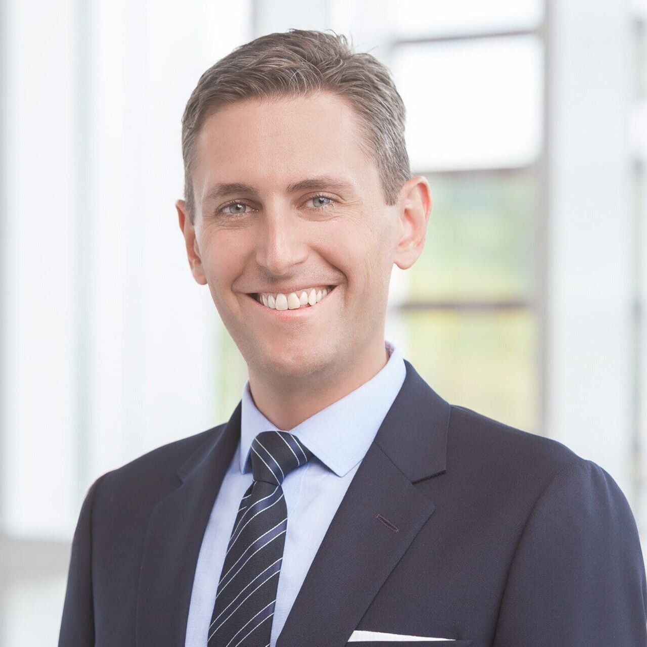 A man in a suit and tie smiling in front of a window for his headshot.