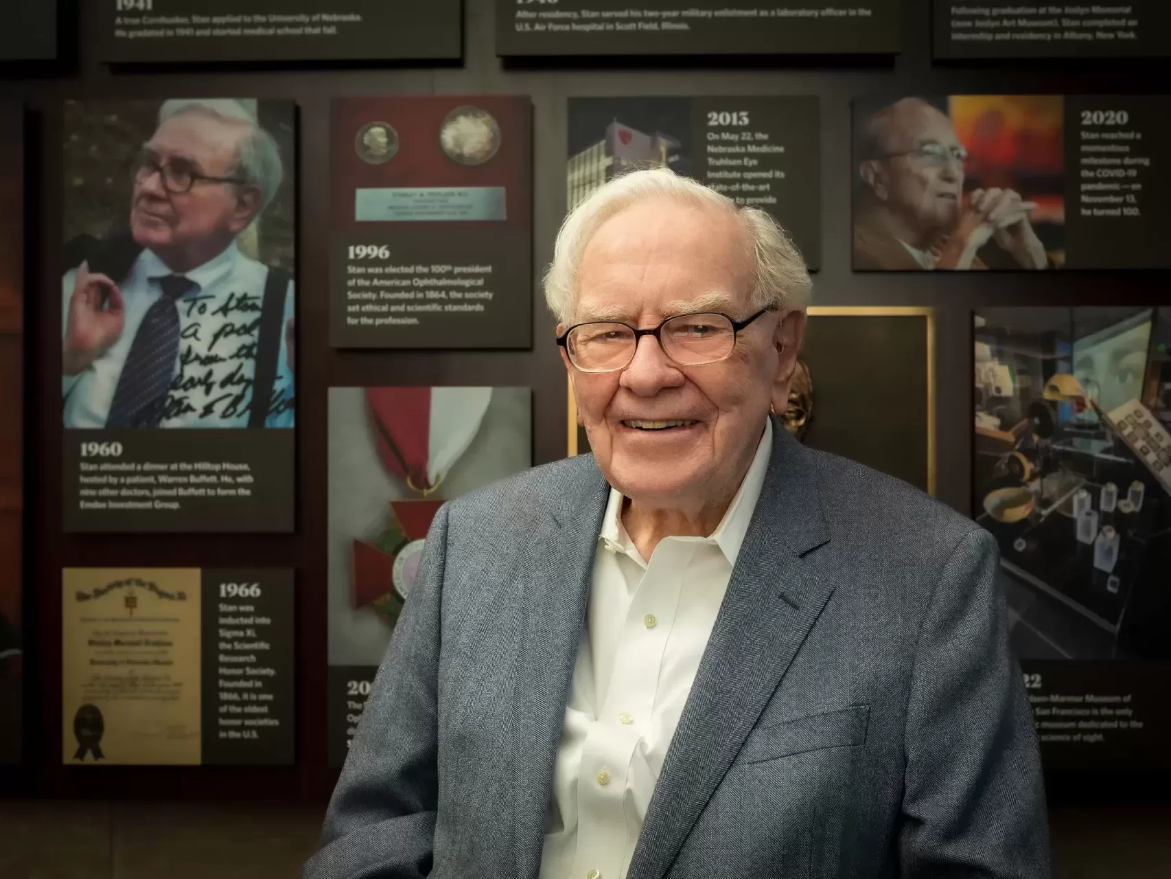 Warren Buffett smiling in front of his biographical photo wall display.