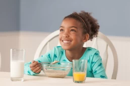 A young girl smiling at a table with a bowl of cereal and orange juice in a food 
photography session.