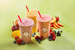 Three colorful smoothie cups with red straws are surrounded by assorted fruits on a light green background.