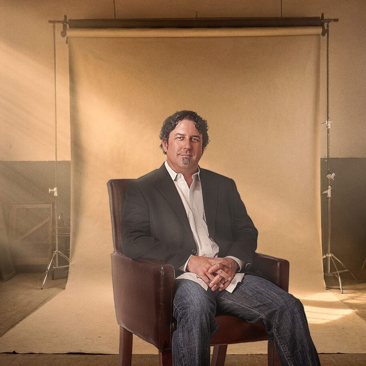 A man sits in a brown armchair for a studio portrait with warm backlighting.