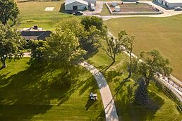 Aerial view of a rural property with a curving driveway, lush lawn, mature trees, and farm outbuildings.