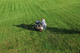 Aerial view of a person mowing a large lawn with a ride-on mower on striped green grass.